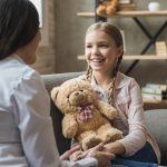 Young, happy child holding a teddy bear whilst talking to her Foster Carer sat on the sofa at home.